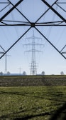 An expansive landscape with several electricity pylons extending across a green field. The pylons stand tall and are silhouetted against a clear blue sky, framed by the metal structure of another pylon in the foreground. Sparse trees are visible in the distance along the horizon.