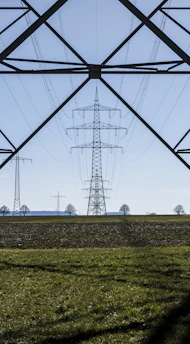 An expansive landscape with several electricity pylons extending across a green field. The pylons stand tall and are silhouetted against a clear blue sky, framed by the metal structure of another pylon in the foreground. Sparse trees are visible in the distance along the horizon.