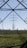 An expansive landscape with several electricity pylons extending across a green field. The pylons stand tall and are silhouetted against a clear blue sky, framed by the metal structure of another pylon in the foreground. Sparse trees are visible in the distance along the horizon.