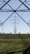 An expansive landscape with several electricity pylons extending across a green field. The pylons stand tall and are silhouetted against a clear blue sky, framed by the metal structure of another pylon in the foreground. Sparse trees are visible in the distance along the horizon.