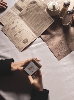 A person is holding a smartphone with a colorful block game on the screen, at a table with printed menus and a set of condiments including a teapot, salt, and pepper shakers. The table has a minimalist design with a festive table mat.