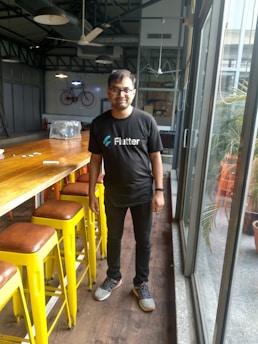 A person wearing a black Flutter t-shirt is standing inside a modern, industrial-style room. The room features high ceilings with exposed ductwork and ceiling fans. There is a long wooden table lined with bright yellow stools. Large glass windows allow natural light to enter, and a bicycle is mounted on the wall as decoration.