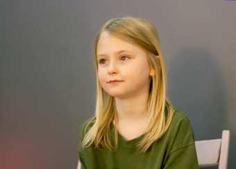 A young child with long blonde hair sits on a chair, wearing a green shirt. The background is a plain gray wall, creating a simple and neutral setting. The child is looking to the side with a serene expression.