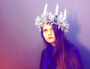 A young girl with long brown hair wears a shiny crown adorned with four white candles. She is dressed in a deep blue cloak, and her expression is calm and thoughtful. The background is a plain muted purple, creating a simple yet striking contrast with her attire.