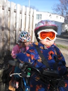 Two children wearing helmets and warm clothing are sitting on a bicycle. The child in the foreground is wearing a colorful snowsuit and orange goggles, while the child in the background is in a pink outfit, also with a helmet. A wooden fence and a residential building are visible in the background under a clear blue sky.
