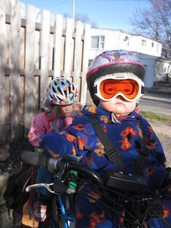 Two children wearing helmets and warm clothing are sitting on a bicycle. The child in the foreground is wearing a colorful snowsuit and orange goggles, while the child in the background is in a pink outfit, also with a helmet. A wooden fence and a residential building are visible in the background under a clear blue sky.
