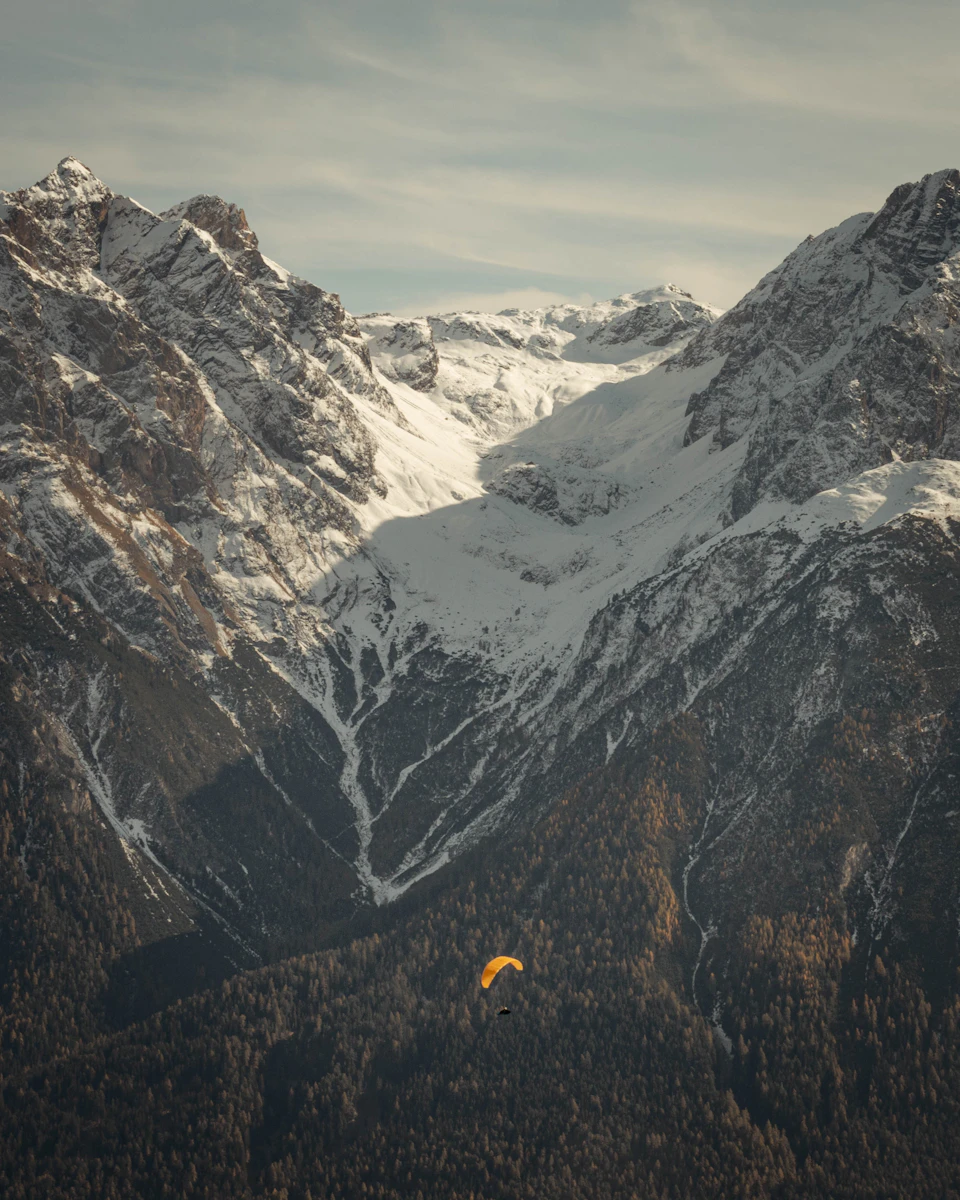 The image depicts a serene mountain landscape with snow-capped peaks and a valley below. The mountains are covered in a blanket of white snow, creating a stunning contrast against the clear blue sky above. In the foreground, there is an orange parachute soaring through the air, adding a vibrant pop of color to the otherwise monochrome scene.