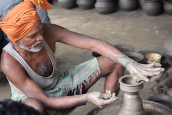 A man is focused on shaping clay on a pottery wheel. He is wearing a bright orange turban and a traditional sleeveless vest. His hands are covered in clay as he carefully molds a pot. Finished pots are visible in the background.