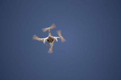 A quadcopter drone flying in a clear blue sky with its propellers spinning visibly. The drone has a white body and is positioned centrally against the expansive sky, capturing a moment of flight.