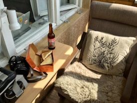 A cozy corner features a plush chair with a botanical print cushion, a small wooden side table with a bottle of beer labeled 'Jaws APA' and a box containing sandwiches. Next to the table, there is a camera bag and a camera with a strap. The area is well-lit by natural light from a nearby window with several small candles on the ledge.