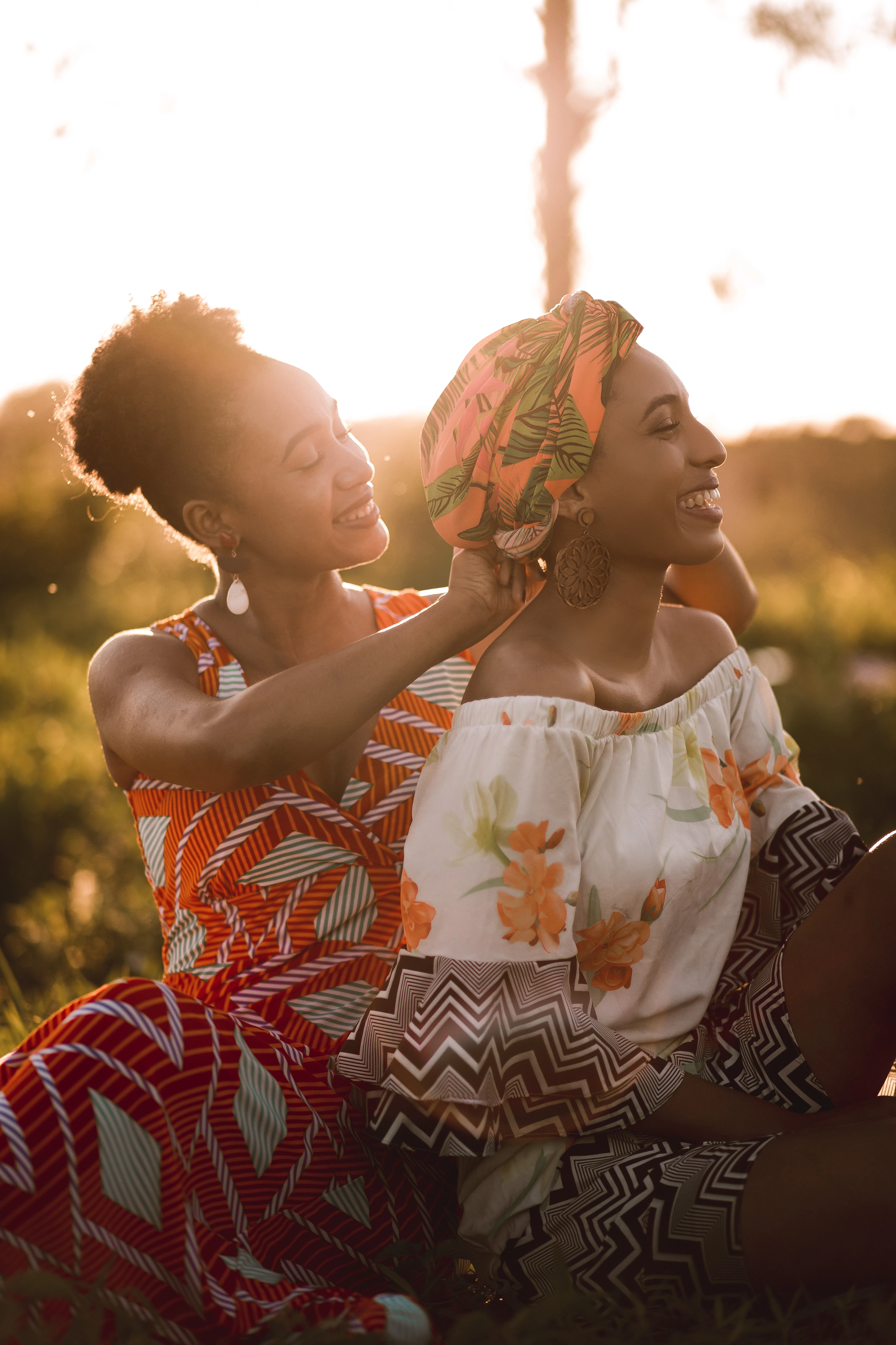 Two women standing close together outdoors in late afternoon light, one adjusting the other's patterned head wrap