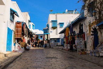 A narrow cobblestone street flanked by white-washed buildings with vibrant blue doors and windows. Various colorful textiles, ceramics, and souvenirs are displayed on the walls and stands, suggesting a lively market atmosphere. A group of people is seen walking along the street, adding to the bustling ambiance.