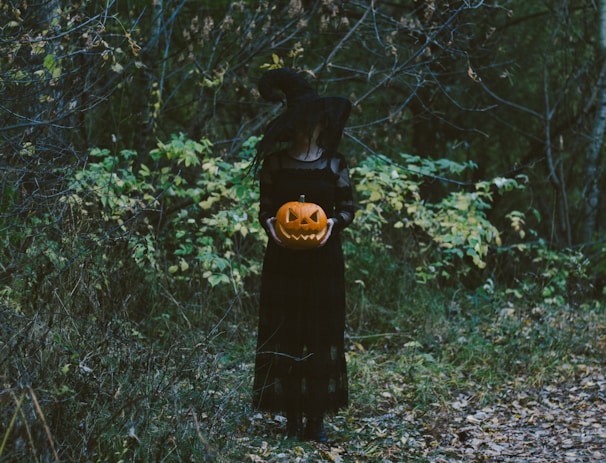 A person dressed as a witch is standing in a dark forest, holding a carved jack-o'-lantern. The witch is wearing a black dress and a pointed hat. The background is filled with dense foliage and trees, creating a mysterious and eerie atmosphere.