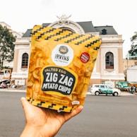 A hand holding a yellow bag of Irvin's Salted Egg Zig Zag Potato Chips with a historic building in the background. The package features a zigzag pattern and a label indicating the chips are dangerously addictive.