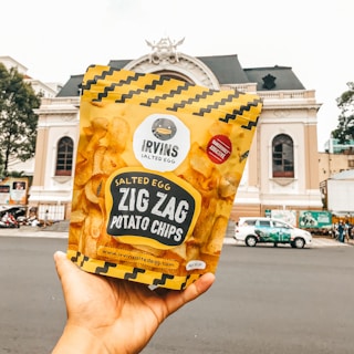 A hand holding a yellow bag of Irvin's Salted Egg Zig Zag Potato Chips with a historic building in the background. The package features a zigzag pattern and a label indicating the chips are dangerously addictive.
