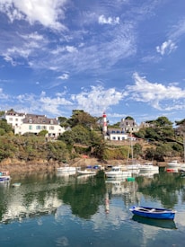 A picturesque coastal scene with several small boats anchored in calm water reflecting a clear blue sky scattered with clouds. In the background, a charming village with houses featuring white walls and slate roofs is nestled among green trees and rocks. A red and white striped lighthouse stands prominently on a hill.