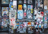 A vibrant collage of street art covers a wall with a mixture of posters, stickers, and graffiti. Various styles of art including abstract shapes, text-based posters, and illustrations of people and animals intermingle. Prominent phrases like 'Life Isn't Beautiful' and 'You Are Free' are visible among the layered visuals.