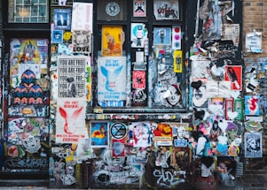 A vibrant collage of street art covers a wall with a mixture of posters, stickers, and graffiti. Various styles of art including abstract shapes, text-based posters, and illustrations of people and animals intermingle. Prominent phrases like 'Life Isn't Beautiful' and 'You Are Free' are visible among the layered visuals.