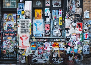 A vibrant collage of street art covers a wall with a mixture of posters, stickers, and graffiti. Various styles of art including abstract shapes, text-based posters, and illustrations of people and animals intermingle. Prominent phrases like 'Life Isn't Beautiful' and 'You Are Free' are visible among the layered visuals.