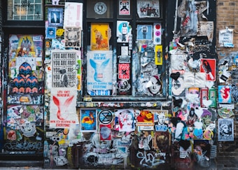 A vibrant collage of street art covers a wall with a mixture of posters, stickers, and graffiti. Various styles of art including abstract shapes, text-based posters, and illustrations of people and animals intermingle. Prominent phrases like 'Life Isn't Beautiful' and 'You Are Free' are visible among the layered visuals.