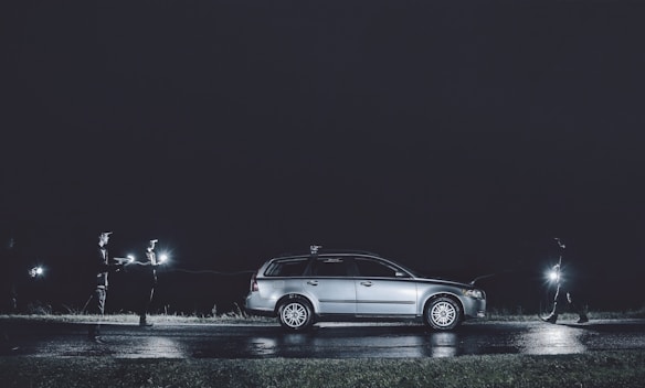 A silver station wagon is parked at night on a wet road, illuminated by the headlights. Three people are positioned around the car, each holding a bright flashlight that casts beams in the darkness. The scene is dimly lit, creating a mysterious and tense ambience.