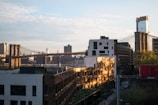 A cityscape featuring the iconic Brooklyn Bridge spanning across a river with urban buildings and skyscrapers in the background. The sunlight creates a warm glow on the structures, casting shadows and highlighting the textures of the architecture. The scene captures a mix of historic and modern elements, with the bridge's cables and towers prominently displayed.