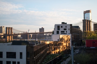 A cityscape featuring the iconic Brooklyn Bridge spanning across a river with urban buildings and skyscrapers in the background. The sunlight creates a warm glow on the structures, casting shadows and highlighting the textures of the architecture. The scene captures a mix of historic and modern elements, with the bridge's cables and towers prominently displayed.