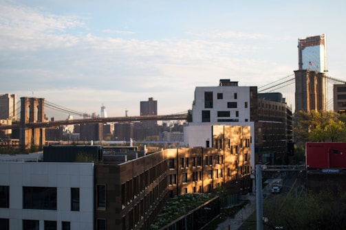 A cityscape featuring the iconic Brooklyn Bridge spanning across a river with urban buildings and skyscrapers in the background. The sunlight creates a warm glow on the structures, casting shadows and highlighting the textures of the architecture. The scene captures a mix of historic and modern elements, with the bridge's cables and towers prominently displayed.