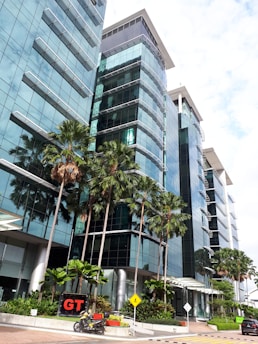 A modern glass building with reflective windows surrounded by tall palm trees. A motorcycle is parked in the foreground near a sign with the letters 'GT'. The sky is partly cloudy, casting a bright light over the scene.