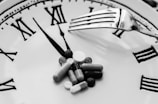 A collection of various pills is arranged on the face of a clock, with a fork resting over them. The clock features Roman numerals and is set to around five minutes past ten.