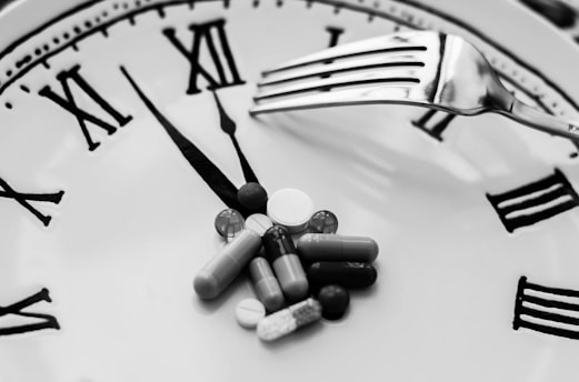 A collection of various pills is arranged on the face of a clock, with a fork resting over them. The clock features Roman numerals and is set to around five minutes past ten.