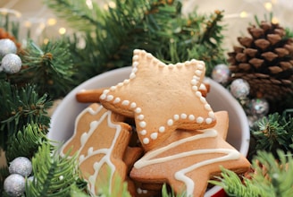 A bowl filled with gingerbread cookies in various shapes, such as stars and leaves, adorned with intricate white icing patterns. Surrounding the bowl are green pine branches, pinecones, and silver ornaments, evoking a festive holiday atmosphere.
