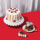 A selection of cakes and desserts is arranged on a bright red tablecloth. A large cake with white frosting, strawberries, and cherry decorations sits on a white stand. Nearby, a smaller chocolate bundt cake topped with leaves and berries is placed on a white dish with a golden spoon. A slice of cheesecake garnished with blueberries and berry syrup sits on the table.