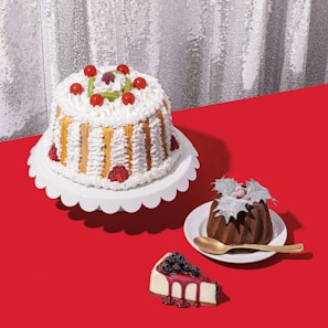 A selection of cakes and desserts is arranged on a bright red tablecloth. A large cake with white frosting, strawberries, and cherry decorations sits on a white stand. Nearby, a smaller chocolate bundt cake topped with leaves and berries is placed on a white dish with a golden spoon. A slice of cheesecake garnished with blueberries and berry syrup sits on the table.