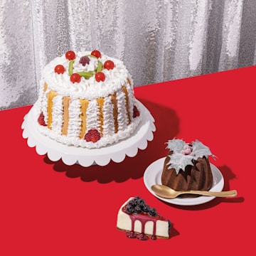 A selection of cakes and desserts is arranged on a bright red tablecloth. A large cake with white frosting, strawberries, and cherry decorations sits on a white stand. Nearby, a smaller chocolate bundt cake topped with leaves and berries is placed on a white dish with a golden spoon. A slice of cheesecake garnished with blueberries and berry syrup sits on the table.