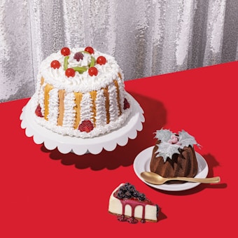 A selection of cakes and desserts is arranged on a bright red tablecloth. A large cake with white frosting, strawberries, and cherry decorations sits on a white stand. Nearby, a smaller chocolate bundt cake topped with leaves and berries is placed on a white dish with a golden spoon. A slice of cheesecake garnished with blueberries and berry syrup sits on the table.