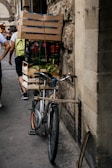 A bicycle is parked against a stone wall, loaded with wooden crates filled with various goods including vegetables. The setting appears to be a bustling street with people walking around, some carrying bags or wearing backpacks. The architecture in the background suggests an old, possibly historic, urban area.
