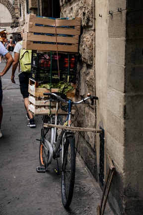 A bicycle is parked against a stone wall, loaded with wooden crates filled with various goods including vegetables. The setting appears to be a bustling street with people walking around, some carrying bags or wearing backpacks. The architecture in the background suggests an old, possibly historic, urban area.