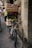 A bicycle is parked against a stone wall, loaded with wooden crates filled with various goods including vegetables. The setting appears to be a bustling street with people walking around, some carrying bags or wearing backpacks. The architecture in the background suggests an old, possibly historic, urban area.