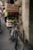 A bicycle is parked against a stone wall, loaded with wooden crates filled with various goods including vegetables. The setting appears to be a bustling street with people walking around, some carrying bags or wearing backpacks. The architecture in the background suggests an old, possibly historic, urban area.