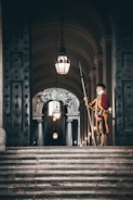 A guard in a colorful uniform stands solemnly with a halberd on a grand staircase. The setting is an ornate hallway adorned with archways and chandeliers. The dim lighting adds a dramatic contrast to the scene, highlighting the intricate details on the walls and ceiling.