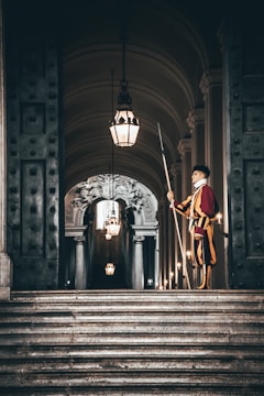A guard in a colorful uniform stands solemnly with a halberd on a grand staircase. The setting is an ornate hallway adorned with archways and chandeliers. The dim lighting adds a dramatic contrast to the scene, highlighting the intricate details on the walls and ceiling.