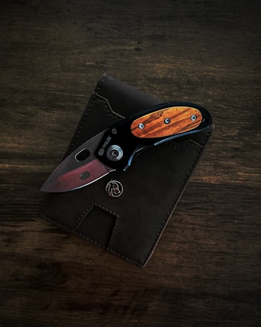 A pocket knife is placed on top of a dark leather wallet. The knife has a wooden handle with metal accents and a visible button or locking mechanism.