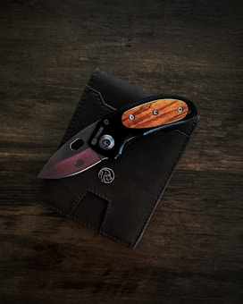 A pocket knife is placed on top of a dark leather wallet. The knife has a wooden handle with metal accents and a visible button or locking mechanism.