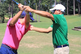 Two individuals are on a golf course. One person in a pink shirt is holding a golf club, while the other in a green shirt appears to be instructing or assisting with the swing. They are surrounded by lush green grass and trees in the background on a sunny day.