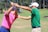 Two individuals are on a golf course. One person in a pink shirt is holding a golf club, while the other in a green shirt appears to be instructing or assisting with the swing. They are surrounded by lush green grass and trees in the background on a sunny day.