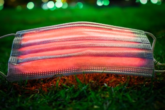 A medical face mask lies on grass, illuminated from below with red lighting. The background features bokeh effects with green and white lights.