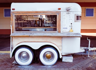 A vintage-style mobile coffee cart with a wood and white metal exterior, featuring a large window displaying coffee-making equipment. Menu and branding are visible on the side, and it is parked on a paved surface.