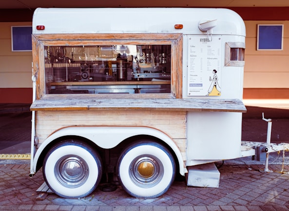 A vintage-style mobile coffee cart with a wood and white metal exterior, featuring a large window displaying coffee-making equipment. Menu and branding are visible on the side, and it is parked on a paved surface.
