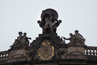 A classical statue features a central figure holding a globe, flanked by two reclining figures on either side. The ornate building facade includes an intricate emblem with a winged creature and shield motif, set against a backdrop of decorative balustrades.